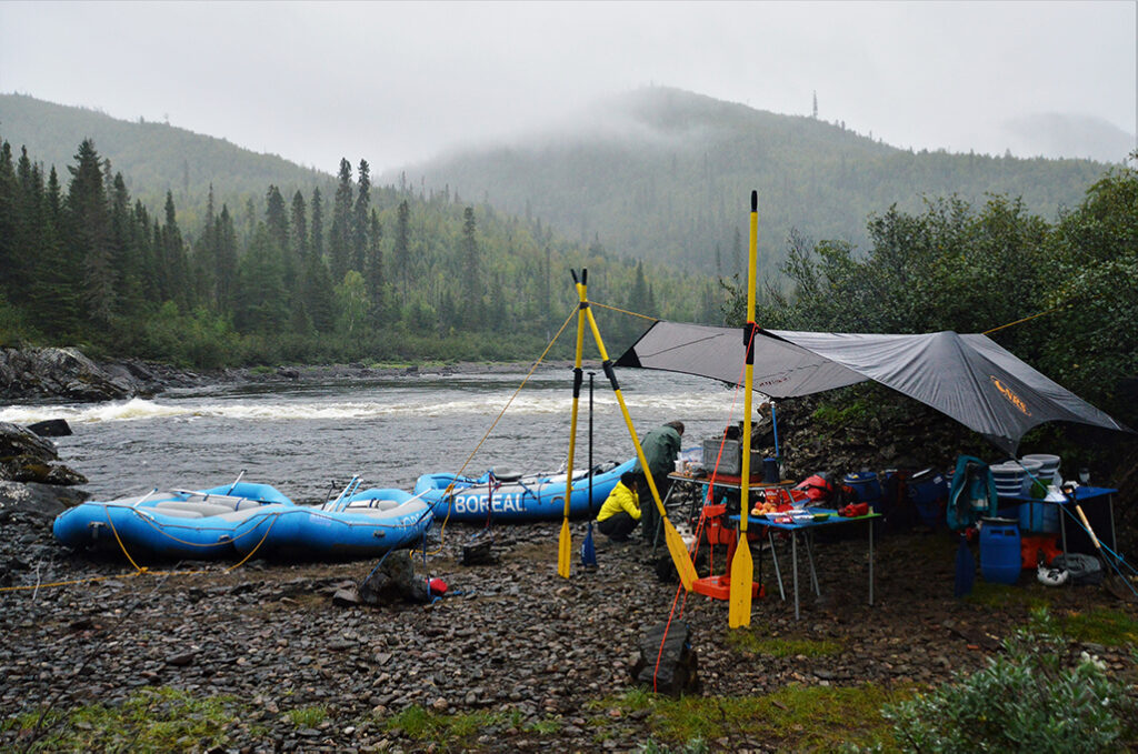 Camp setup on a rainy day. Tarp is held by two paddles for a dry area. 3 rafts pulled on shore along Muteshekau Shipu (Magpie River).